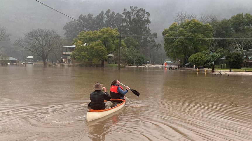 Two people row a canoe in floodwaters in a town.