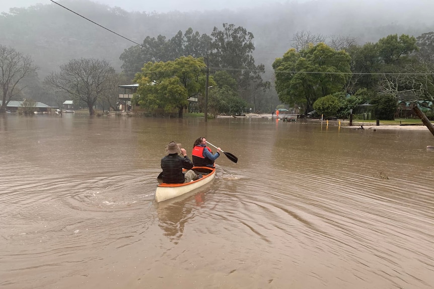 Two people row a canoe in floodwaters in a town.