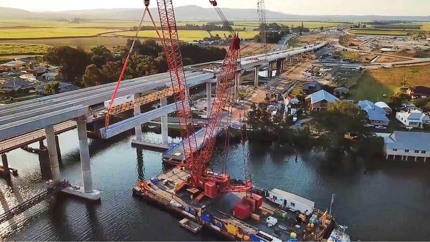An aerial shot of a girder being installed in a concrete bridge by a large red crane over a river.