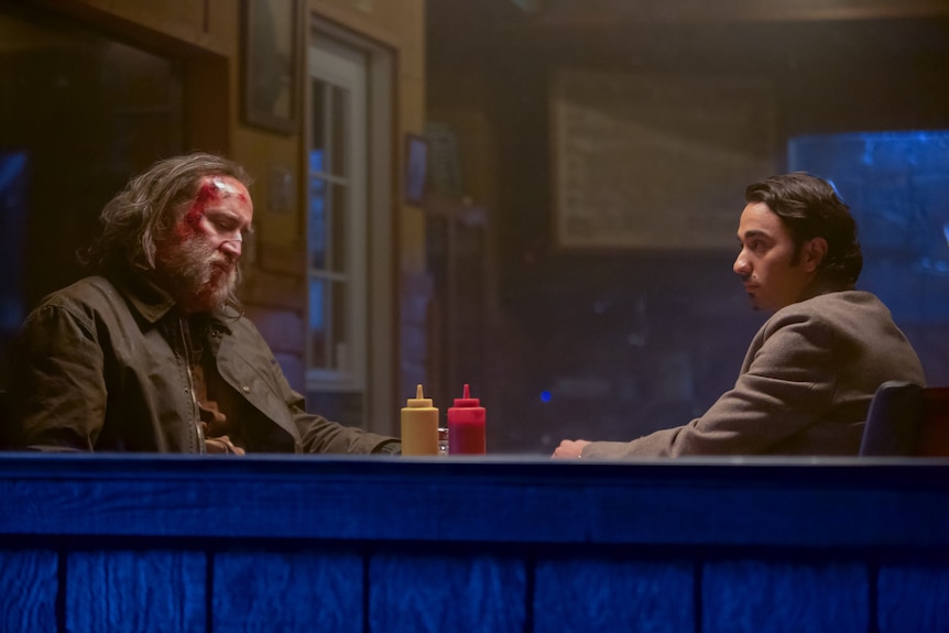 A battered-looking Nicolas Cage with long greying hair sits with a 20-something man with a goatee in a diner