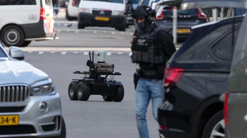 Looking down a street cordoned off with red and white police tape, an autonomous police robot drives between cars and officers.