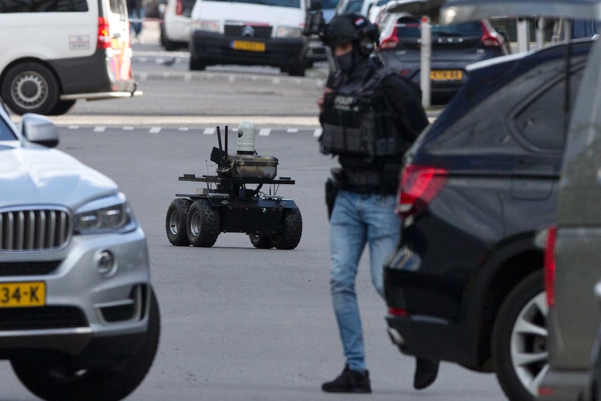 Looking down a street cordoned off with red and white police tape, an autonomous police robot drives between cars and officers.