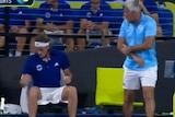 An older man holds his right elbow while like down at a young man sitting on the bench, a broken racquet at his side
