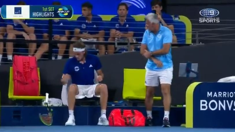 An older man holds his right elbow while like down at a young man sitting on the bench, a broken racquet at his side