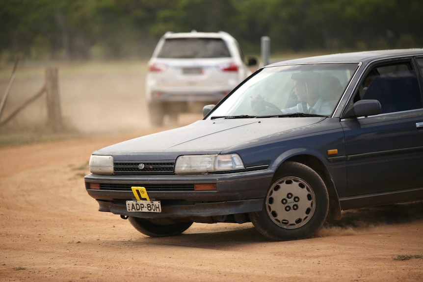 An older model sedan with an L plate displayed on the front pulls out on a dusty dirt road on a farm