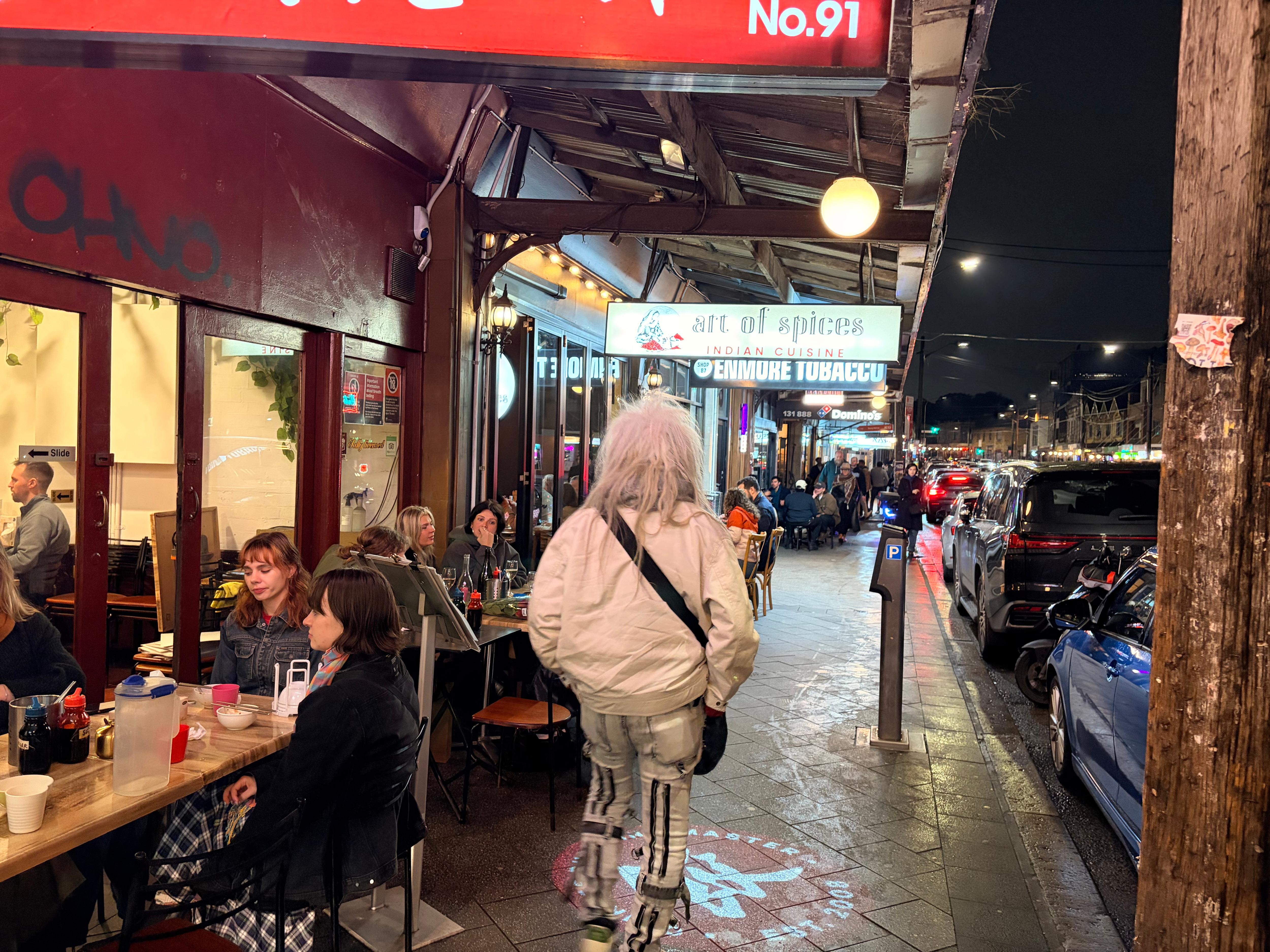 Man with white hair walking as people eat and drink on street at night