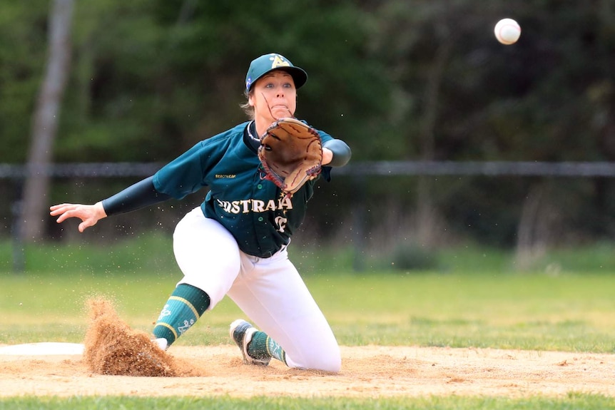 A woman slides on the dirt as she stretches to catch a baseball.