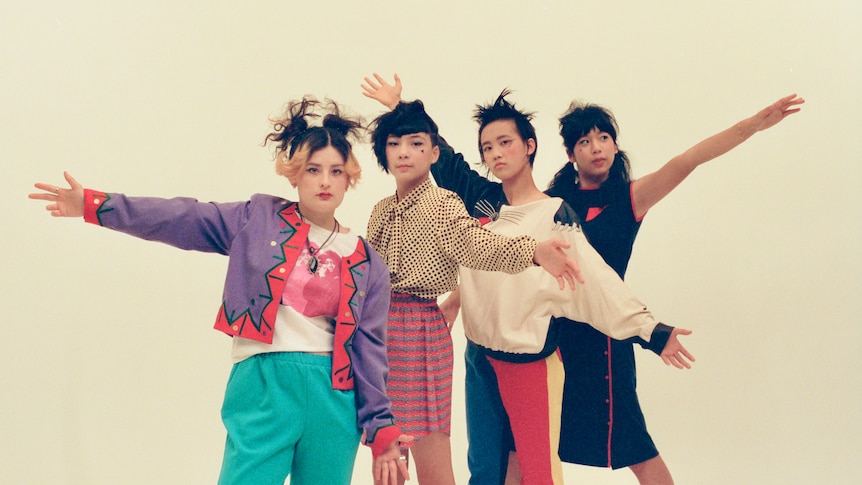Four young women in colourful clothing and spiked hair hold their arms outstretched