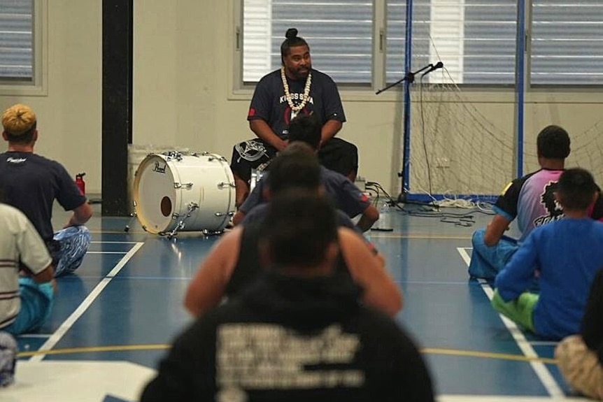 Man seen in distance playing the drums as young Samoan teens sit on the ground, their backs facing us. 