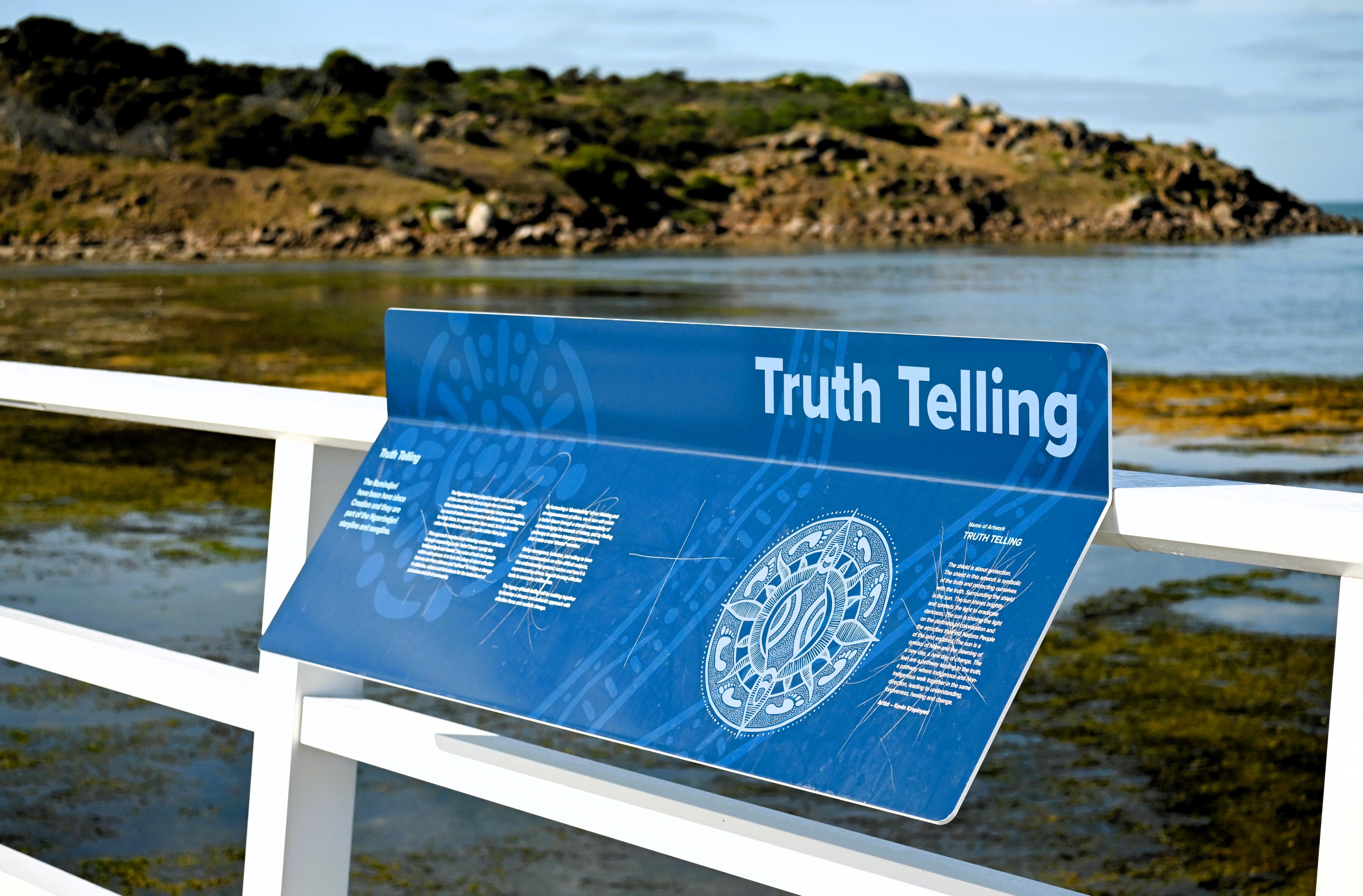 A damaged blue sign with writing and graphics attached to a bridge fence with the sea and an island visible behind