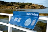 A damaged blue sign with writing and graphics attached to a bridge fence with the sea and an island visible behind