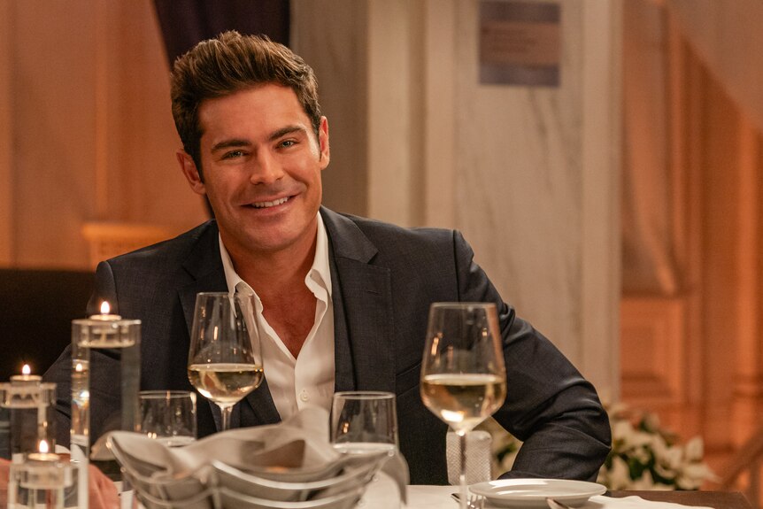 Zac Efron as Chris Cole. He is pictured wearing a suit with glasses of wine on the table in front of him.