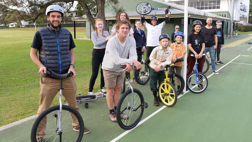 A group of unicycle enthusiasts including adults and young people holding their unicycles at the netball courts, 12 in number