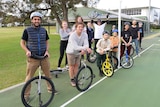 A group of unicycle enthusiasts including adults and young people holding their unicycles at the netball courts, 12 in number