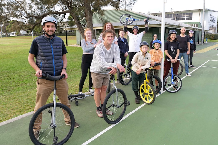 A group of unicycle enthusiasts including adults and young people holding their unicycles at the netball courts, 12 in number