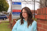 A woman with long brown hair and glasses leans against a brick wall with a hospital sign in the background. 