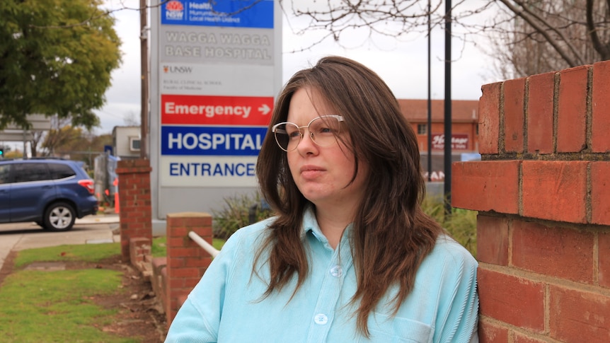A woman with long brown hair and glasses leans against a brick wall with a hospital sign in the background. 