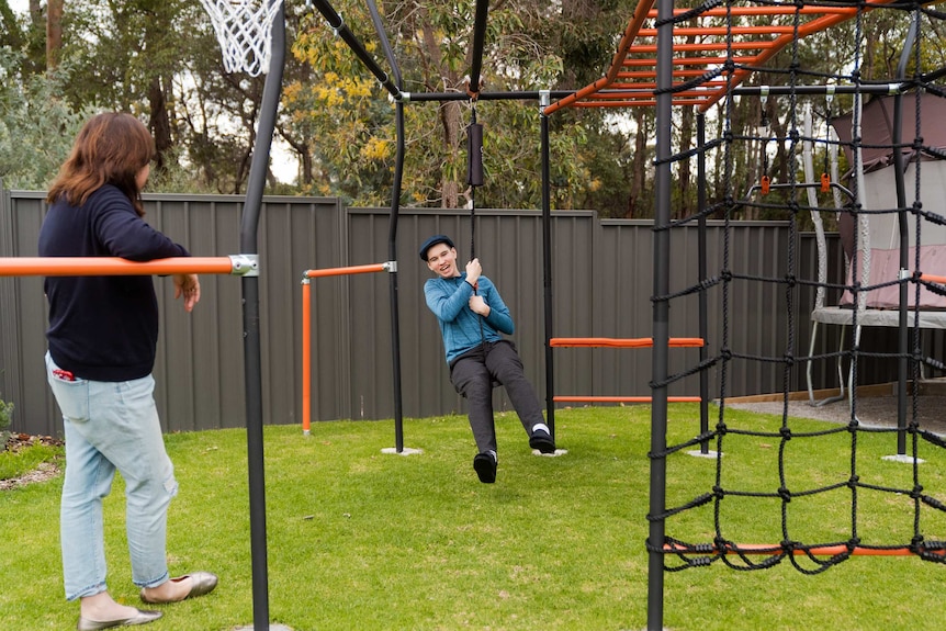 A teenage boy rides on a flying fox in a backyard as his mother watches on.