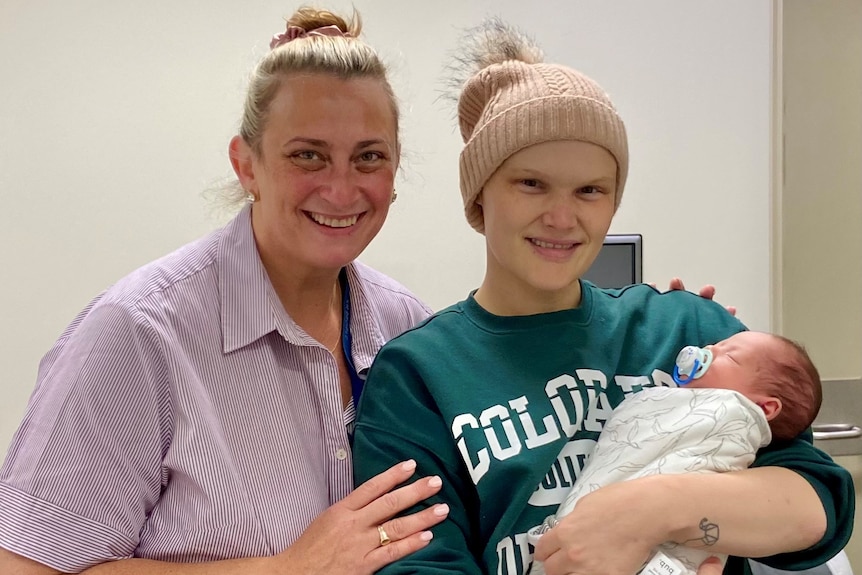 a woman puts her arm arounda  younger woman in a beanie, holding a newborn