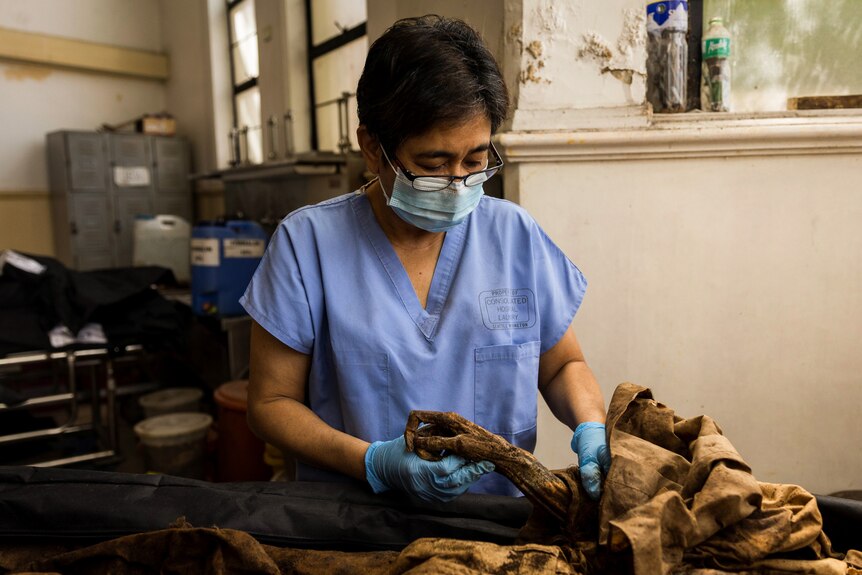 A woman with cropped brown hair wears scrubs, a mask and glasses while inspecting remains.