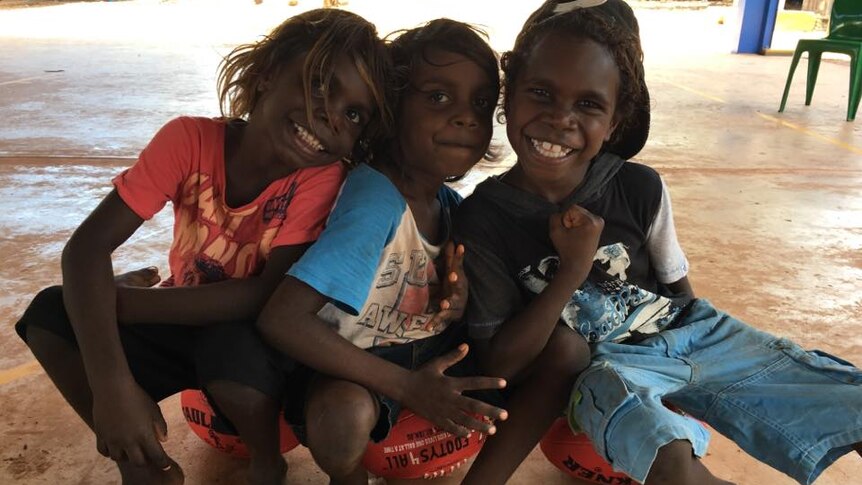 Three young Aboriginal boys smiling and sitting on AFL footballs after a sports clinic.