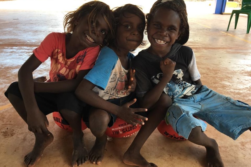 Three young Aboriginal boys smiling and sitting on AFL footballs after a sports clinic.