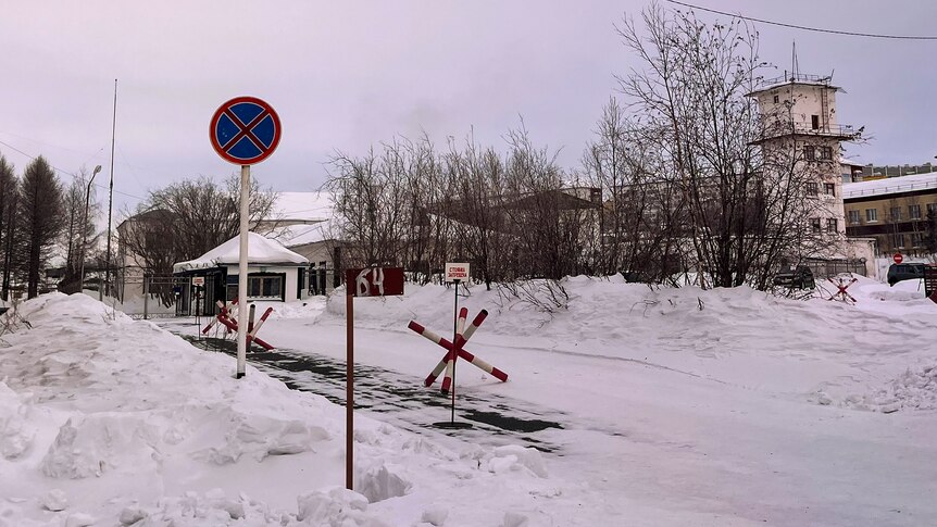 The snow-covered entrance to a Russian prison camp, showing road barriers and guard towers.