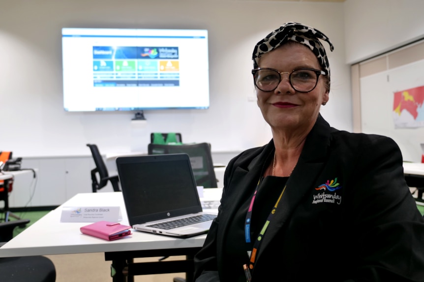 a woman in a corporate black uniform sits in a chair in a large room in front of a laptop looking at the camera