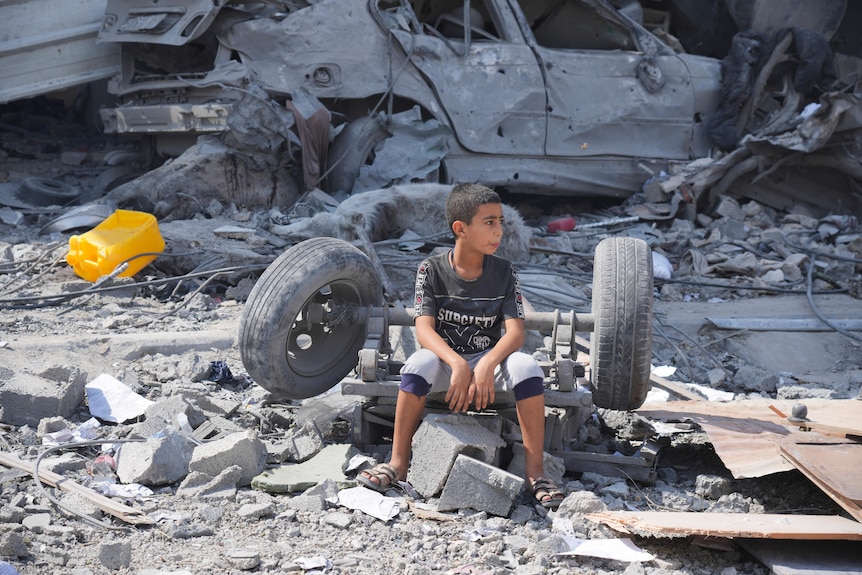 A boy looks to his left sitting on a piece of rubble in a destroyed street