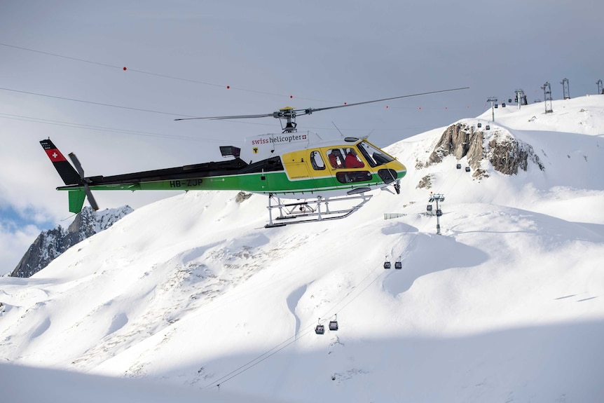 A green and yellow helicopter hovers above the ski slope