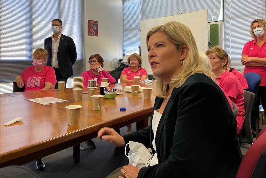 A woman with shoulder-length blonde hair, wearing a black jacket, sitting at a conference table with other people.