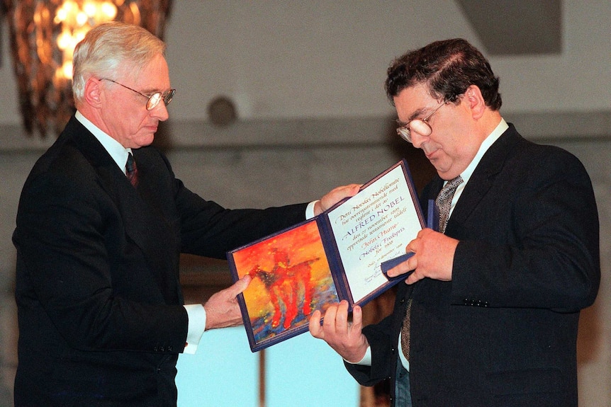 Two men in suits exchange a folder and medal at the Nobel Prize ceremony.