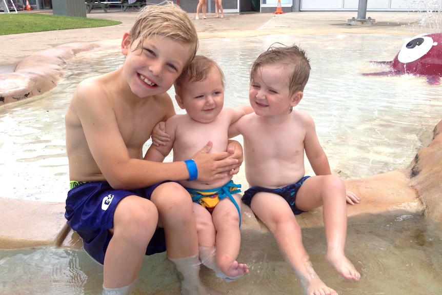 A young boy and two toddlers sit,arms around each other, on the edge of a kiddie pool. They are wearing bathers.
