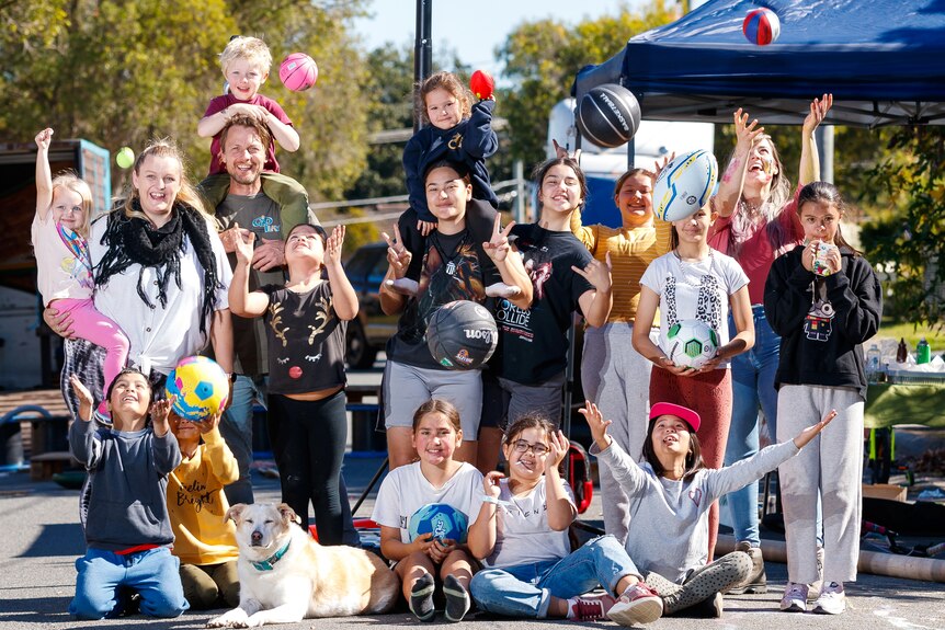 An image of a group of children, teenagers and adults smiling with hands up and balls flying on leafy street in front of gazebo 