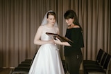 A bride in a white dress and veil talks to a woman dressed in black with a  folder in her hands