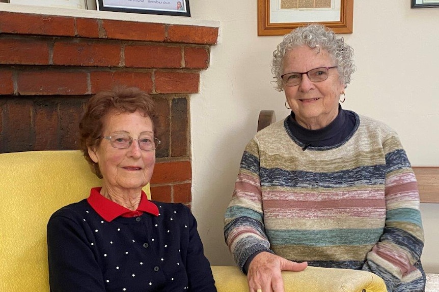 Two smiling older women, one on left has short brown hair, on the right has grey curly hair, both wear glasses. Brick fireplace.