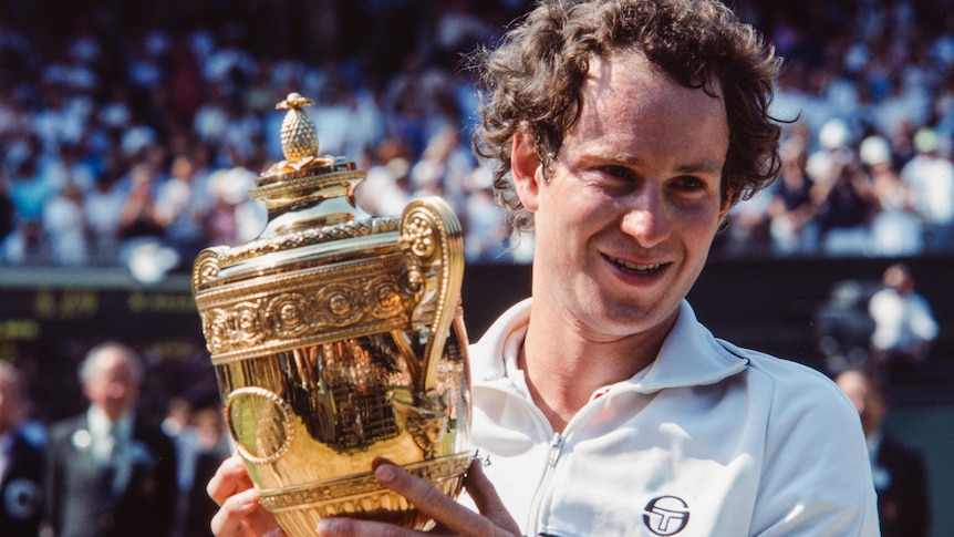 John McEnroe smiles and holds up the Wimbledon trophy