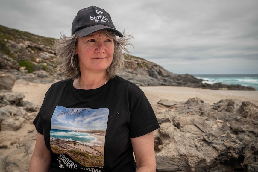 Caroline Paterson sits on a remote beach, wearing a black 'birdlife' cap, flowing grey hair and a black t-shirt.