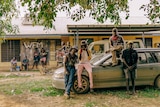 Four members of Wildfire Manwurrk lean up against a silver car. Behind them a group of people hang around outside a building.