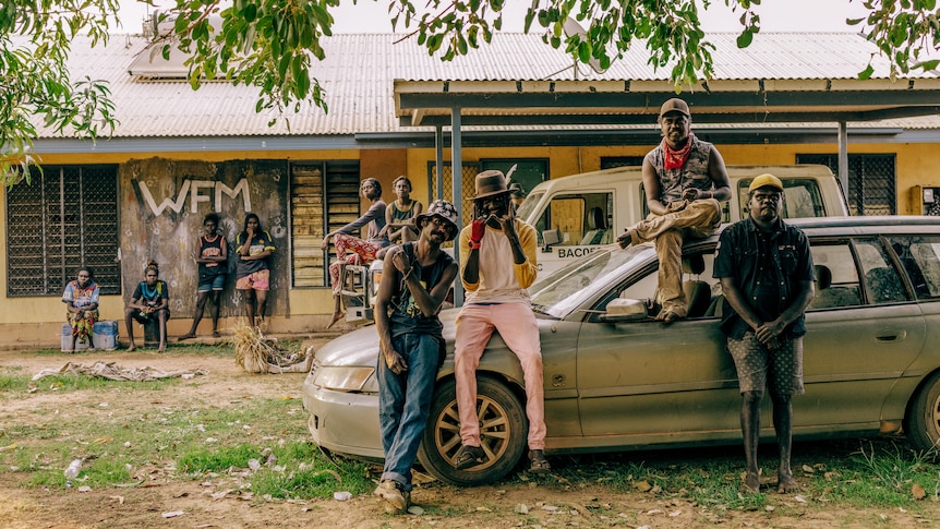 Four members of Wildfire Manwurrk lean up against a silver car. Behind them a group of people hang around outside a building.
