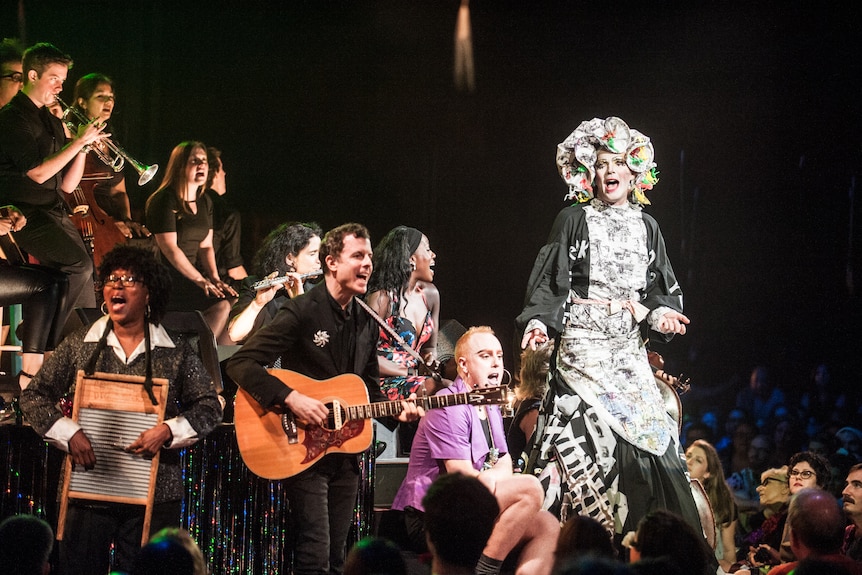 Matt Ray, a white man in a black suit with guitar, & Taylor Mac, a white drag performer, are onstage with singers and musicians