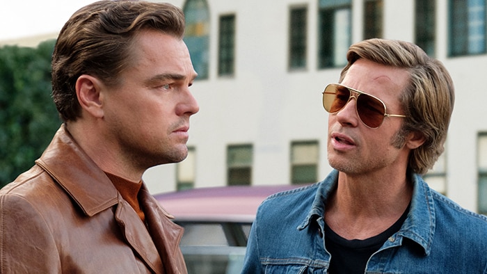 Two men stand in the street, one looks away with serious expression while the man wearing sunglasses faces him and speaks.