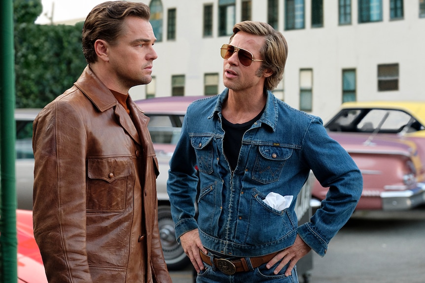 Two men stand in the street, one looks away with serious expression while the man wearing sunglasses faces him and speaks.