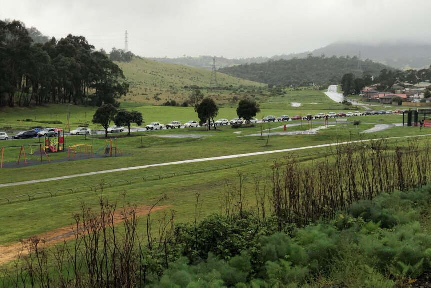 Cars queueing up along a road waiting for a COVID test on an overcast day with green hills in the background.