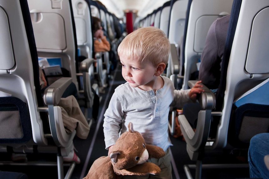 A young child totes a soft toy wikth him as he wanders down the aisle of an aircraft
