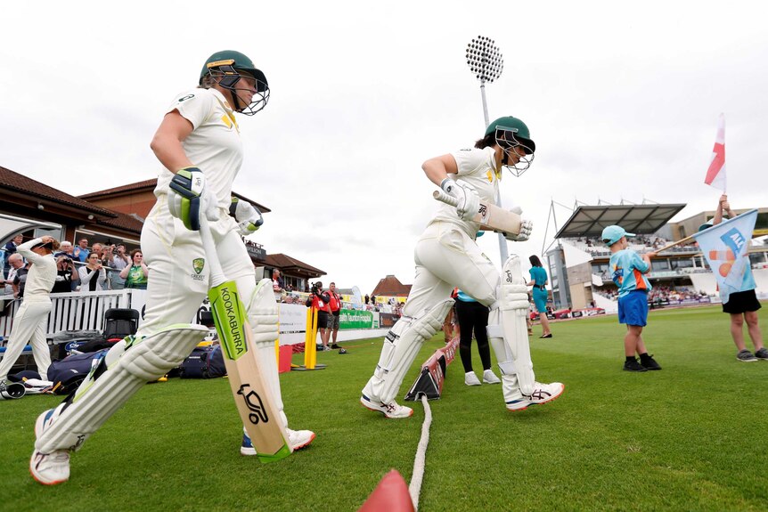 Alyssa Healy and Nicole Bolton, wearing cricket whites and helmets, walk across the rope.