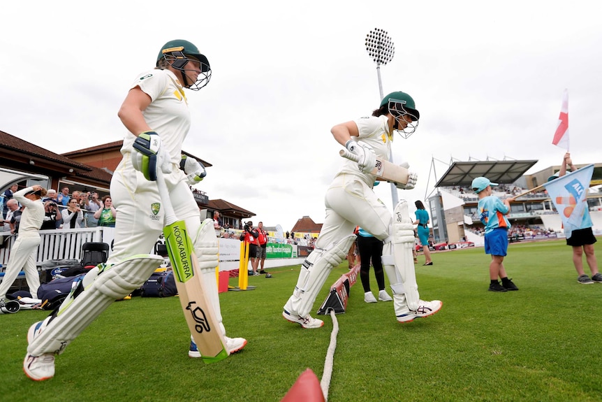 Alyssa Healy and Nicole Bolton, wearing cricket whites and helmets, walk across the rope.