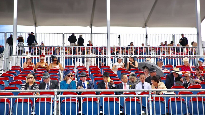 A crowd begins to gather in the stands at the Melbourne Cup with hats and an umbrella.