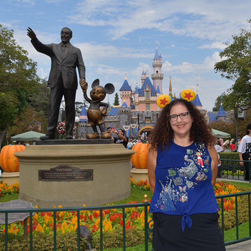 A smiling adult woman wearing Mickey Mouse ears in front of a statue of Walt Disney and Mickey Mouse at a Disney theme park