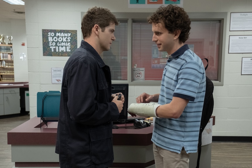 Two young men face each other uncomfortably in a school library, one wearing dark clothing and the other in a cast